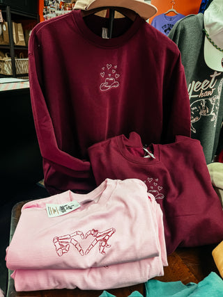 Maroon crewneck and pink tee with Valentine's Day designs by Hayley Skinner on a table in a store.