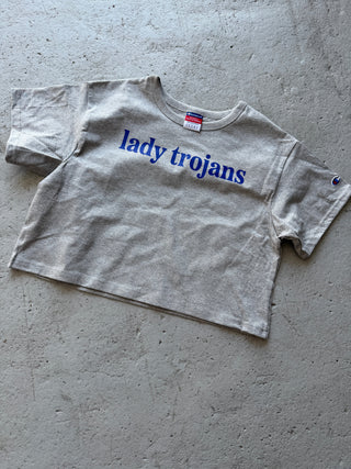 A light gray Champion t-shirt with 'lady trojans' printed in blue text, laid flat on a textured concrete surface.