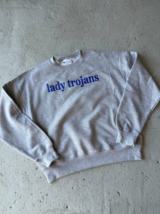 A light heather gray crewneck sweatshirt with 'lady troyans' printed vertically in blue on the chest, laid flat on a textured concrete surface.